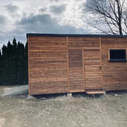 Drewniana konstrukcja z oknem i drzwiami, prawdopodobnie altana, na fundamencie, otoczona zielenią i drzewami pod pochmurnym niebem.