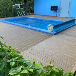 Zadaszony basen z niebieską mozaiką i zwiniętą pokrywą, otoczony drewnianym tarasem i zielenią, z błękitną ścianą budynku w tle i klimatyzatorem na elewacji.