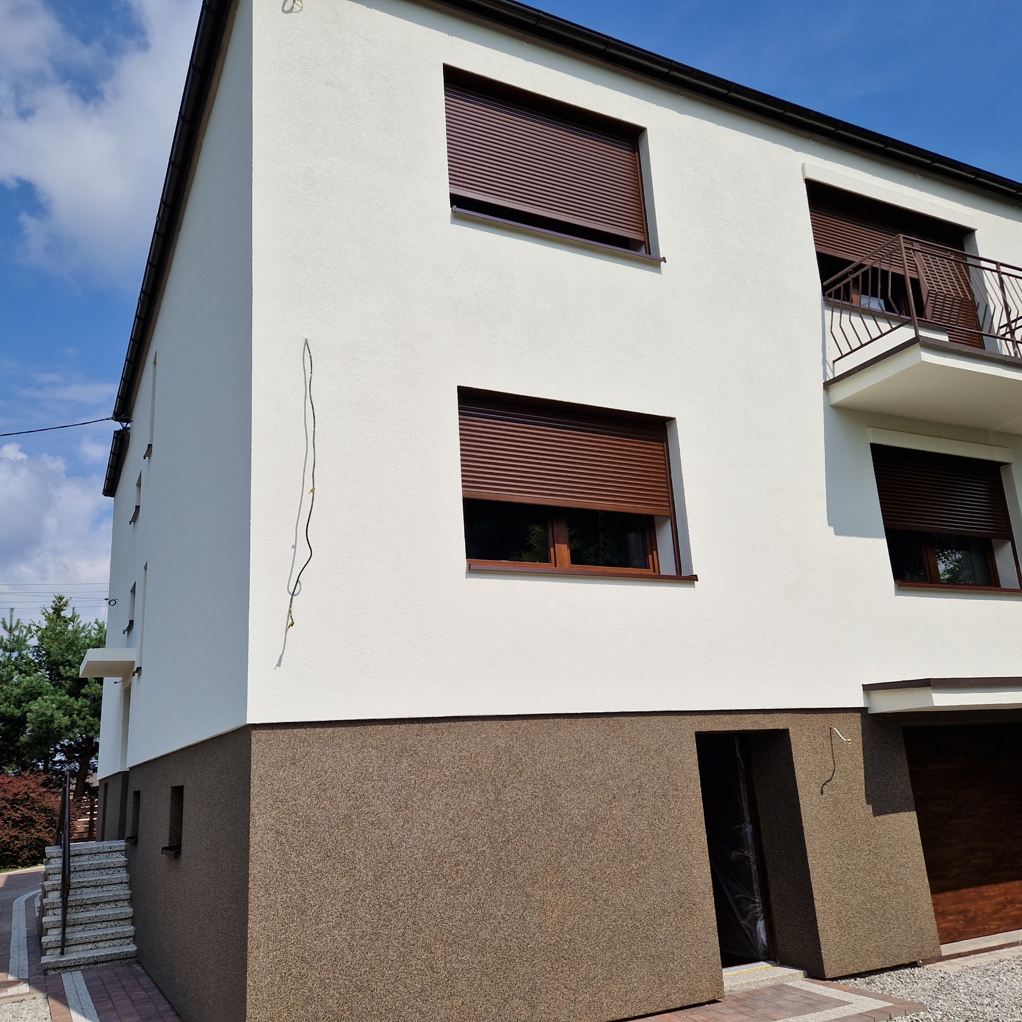 Dwukondygnacyjny dom z białą elewacją górną i brązową elewacją dolną, brązowe rolety w oknach, balkon z balustradą, widok z dołu na bocznej ścianie zwisający kabel.