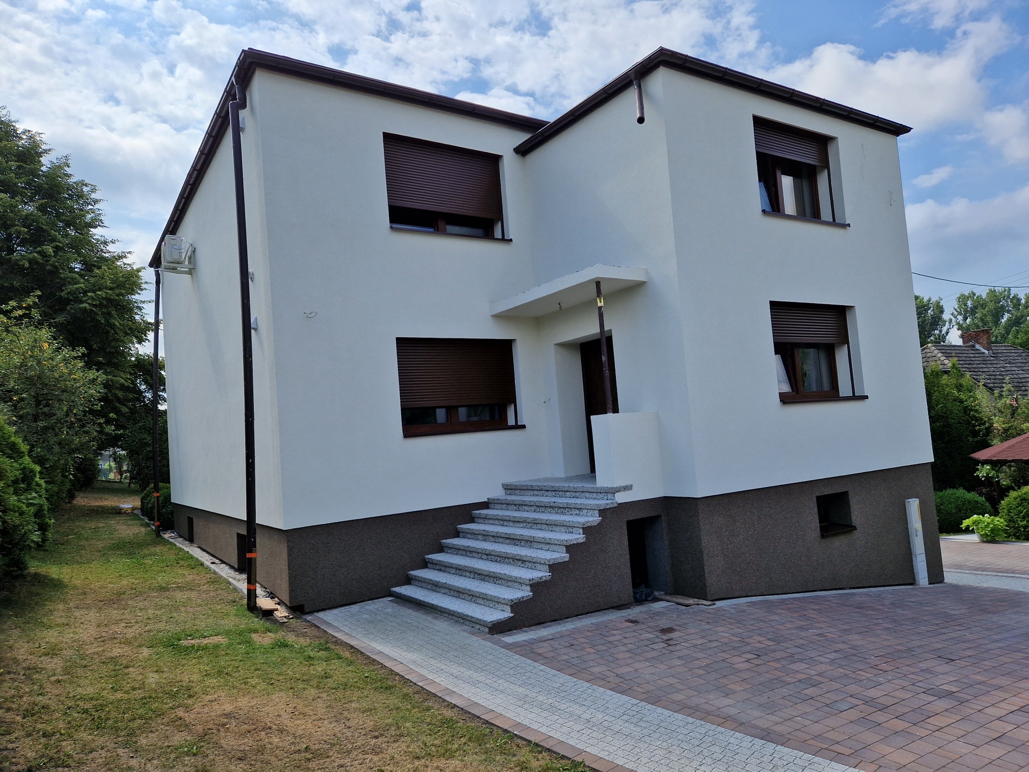 Dwukondygnacyjny dom z jasną elewacją, brązowymi roletami i podmurówką, schody wejściowe wyłożone jasną kostką, widok z zewnątrz w słoneczny dzień.