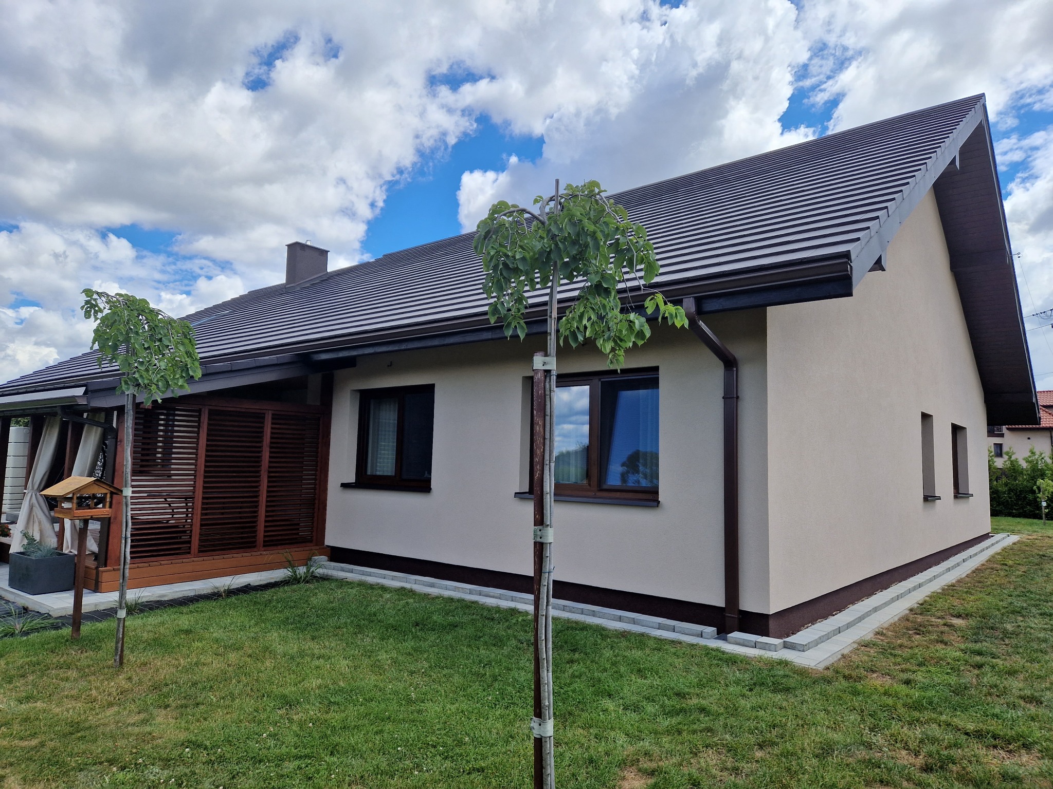 Nowoczesny dom jednorodzinny z beżową elewacją, brązowymi detalami i ciemnym dachem, widok z ogrodu z dwoma młodymi drzewkami na pierwszym planie.