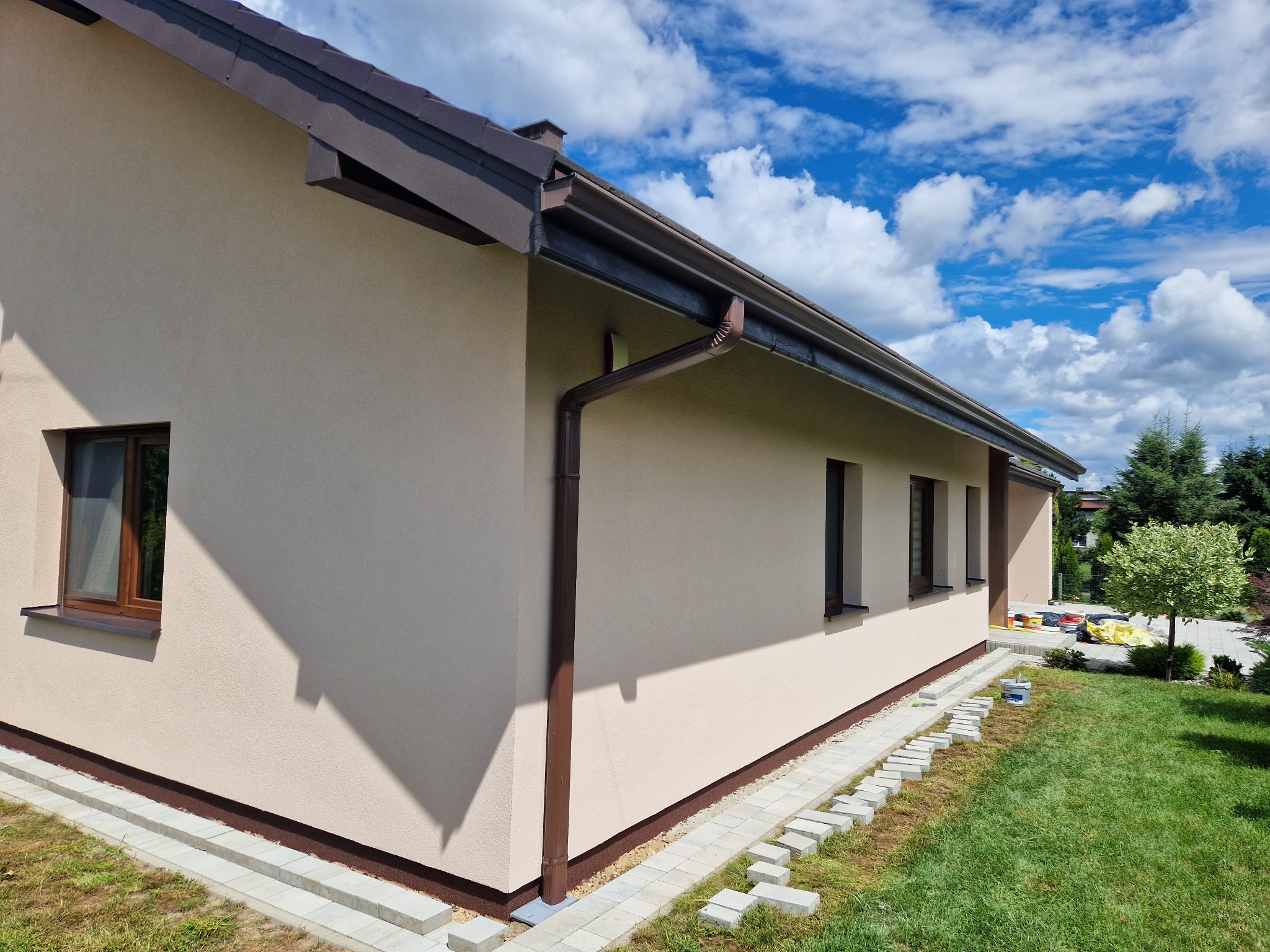 Jasnobeżowa elewacja domu jednorodzinnego z brązowymi oknami i rynnami, widok z boku na tle błękitnego nieba z obłokami, kostka brukowa ułożona wzdłuż ściany.