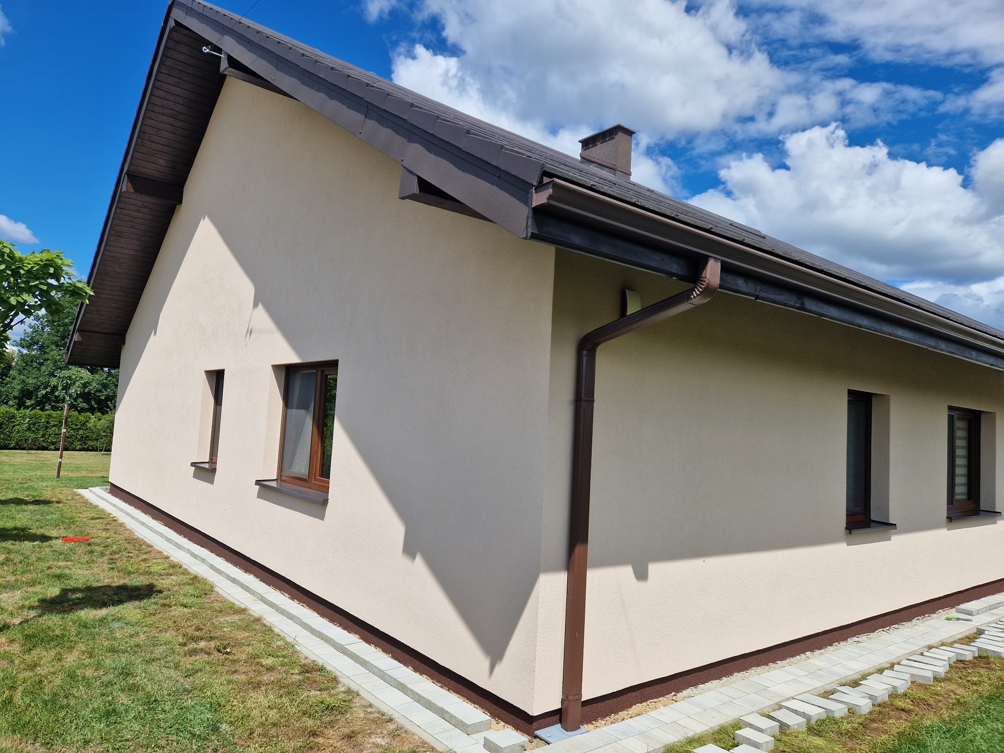 Jasna elewacja domu jednorodzinnego w Strumieniu, z brązowymi oknami i dachem, widok z boku na tle błękitnego nieba z chmurami.