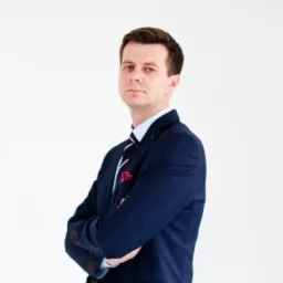 Portret mężczyzny w granatowym garniturze, z rękami skrzyżowanymi na piersiach, na białym tle.