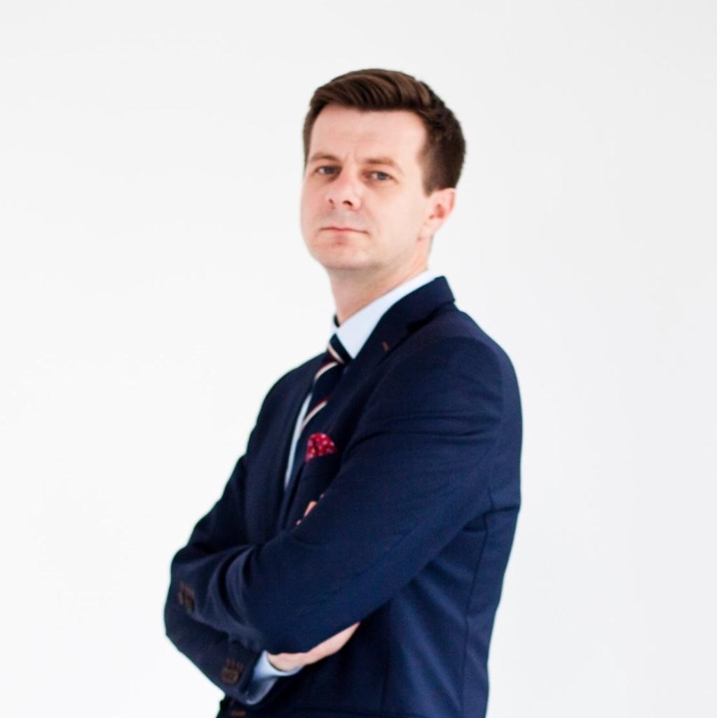 Portret mężczyzny w granatowym garniturze, z rękami skrzyżowanymi na piersiach, na białym tle.