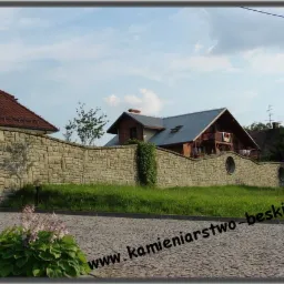 Kamienny mur okalający posesję z domami w tle, widoczny z poziomu brukowanej drogi.