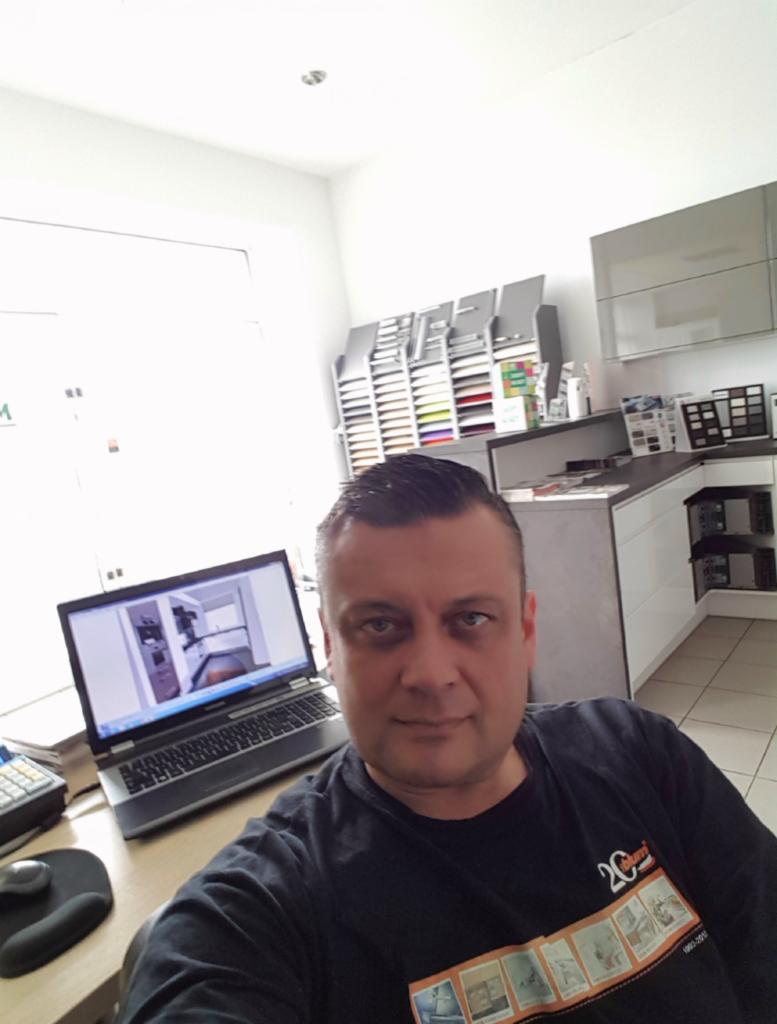 Mężczyzna przy biurku z laptopem prezentującym projekt kuchni na wymiar. W tle ekspozycja próbek materiałów i zabudowa kuchenna.