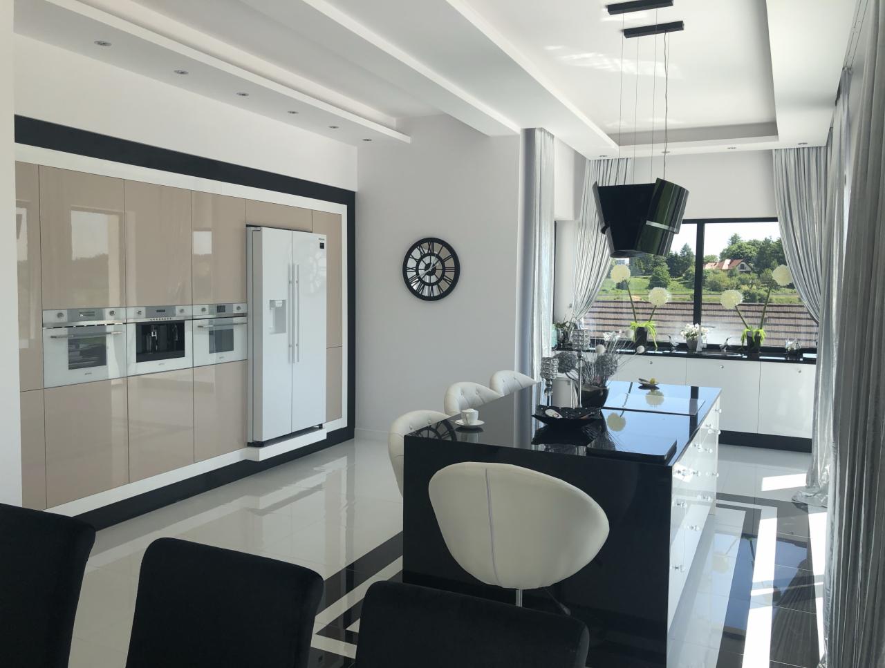 Elegancka kuchnia w nowoczesnym stylu: beżowe szafki z czarnymi obramowaniami, biała lodówka, wbudowane piekarniki, czarny wyspowy blat, białe krzesła, widok na ogród z okna.