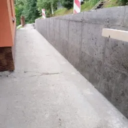 Betonowy mur oporowy wzdłuż drogi, widoczny z poziomu chodnika, z elementami drewnianymi i znakami drogowymi w tle.