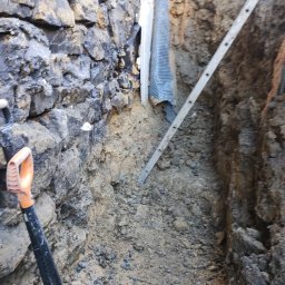 TOMINI Tomasz Smolin - Wykop w ziemi z odsłoniętą kamienną ścianą, widoczna drabina oparta o ścianę wykopu i fragment łopaty z pomarańczowym uchwytem.