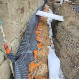 TOMINI Tomasz Smolin - Ocieplenie i wzmocnienie kamiennej ławicy fundamentowej. Widoczna folia kubełkowa, geowłóknina i worki z kamieniem. W tle pracownicy w odblaskowych kamizelkach.