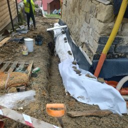 TOMINI Tomasz Smolin - Wykop w ziemi wzdłuż kamiennej ściany fundamentowej, zabezpieczony geowłókniną i folią kubełkową, z widocznymi rurami drenażowymi i pracownikiem w odblaskowej kamizelce w tle.