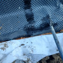 TOMINI Tomasz Smolin - Aplikacja hydroizolacji na folii kubełkowej fundamentu za pomocą pędzla, widoczna geowłóknina i ziemia.