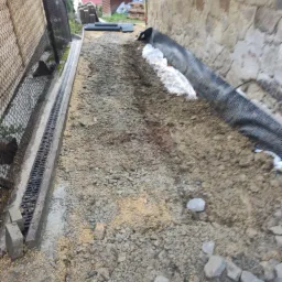 Przygotowanie terenu pod budowę: drenaż liniowy z kratką, warstwa gruntu z drobnym kruszywem, geowłóknina przy kamiennej ścianie, widoczne elementy drenażu i kamienie.