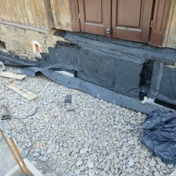 TOMINI Tomasz Smolin - Ocieplenie kamiennej ławicy fundamentowej z użyciem hydroizolacji bitumicznej i folii kubełkowej, widoczne wzmocnienie betonowe, gruz i elementy konstrukcyjne w trakcie prac.