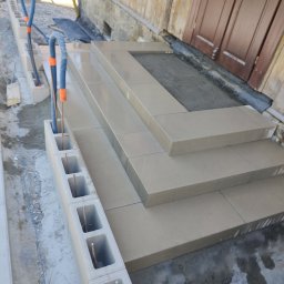 TOMINI Tomasz Smolin - Schody wejściowe wyłożone jasnymi płytami kamiennymi, obok wzniesiona ściana z betonowych pustaków z wystającymi prętami zbrojeniowymi i peszlami instalacyjnymi, widok z góry.