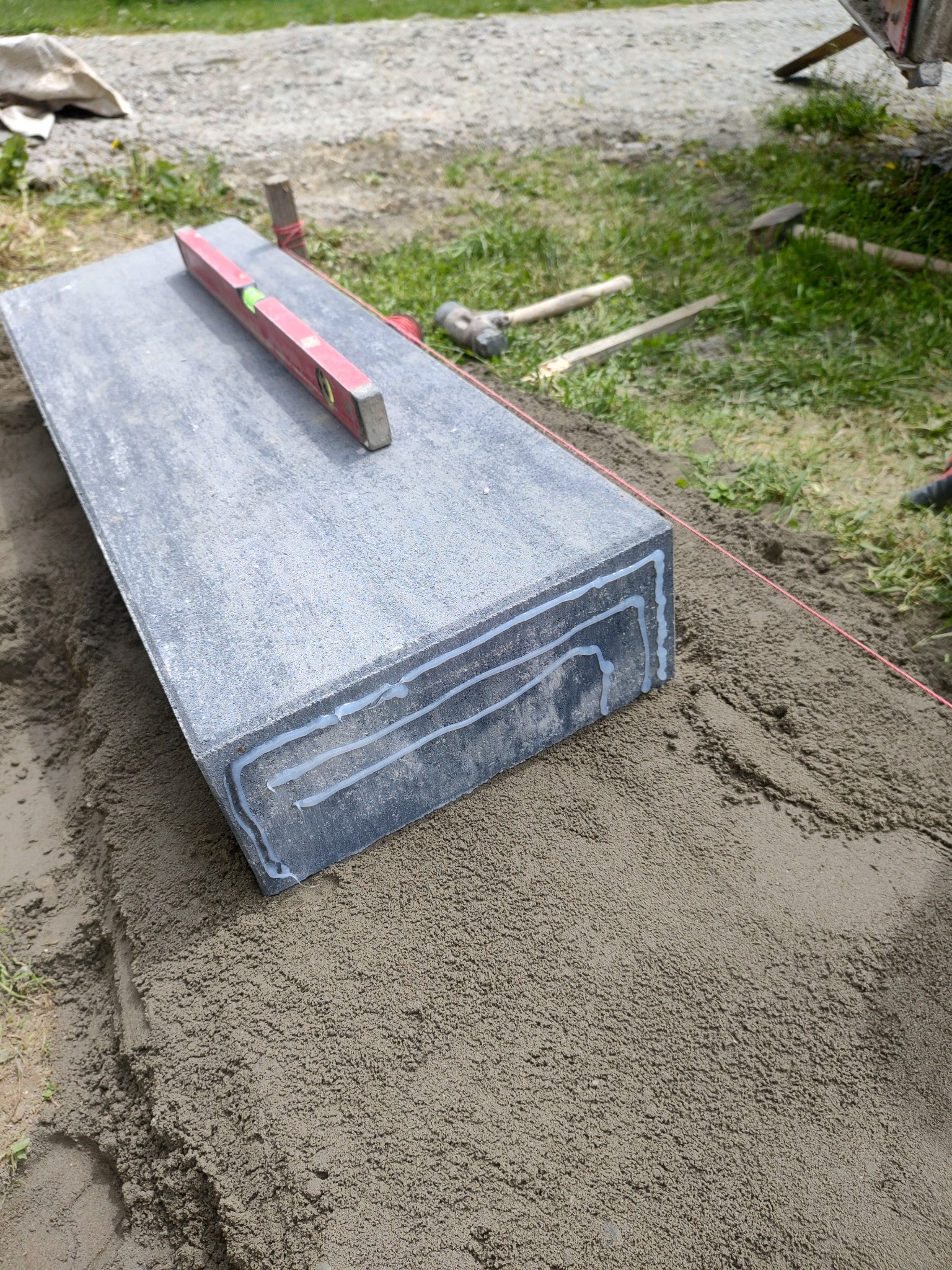 Pojedynczy, duży element betonowy z warstwami kleju na krawędzi, leżący na podsypce piaskowej, poziomowany czerwoną poziomicą, w tle narzędzia brukarskie i trawa.