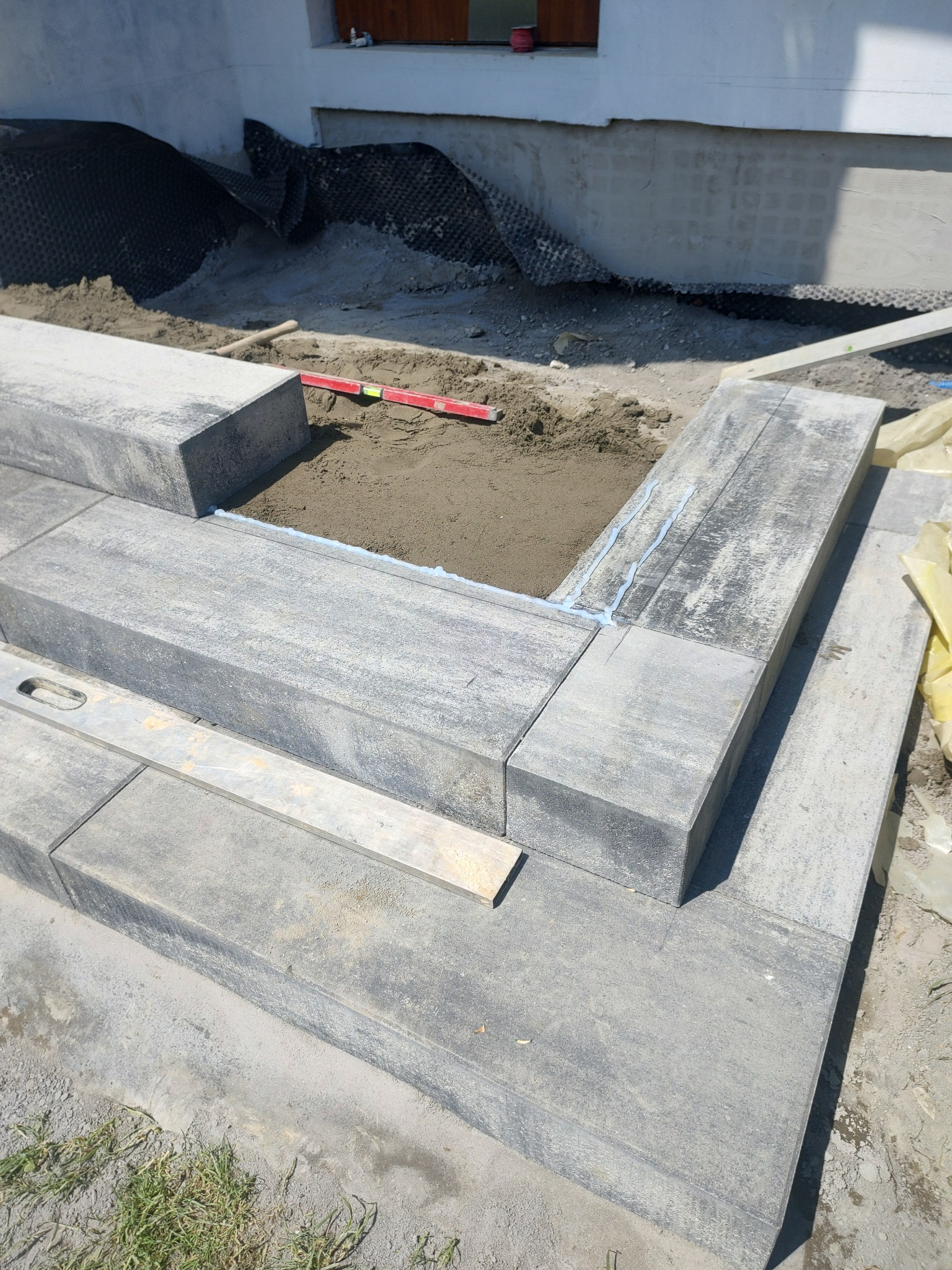 Budowa schodów zewnętrznych z kostki brukowej, widoczny poziomica i zaprawa klejowa, w tle fundament budynku z izolacją.