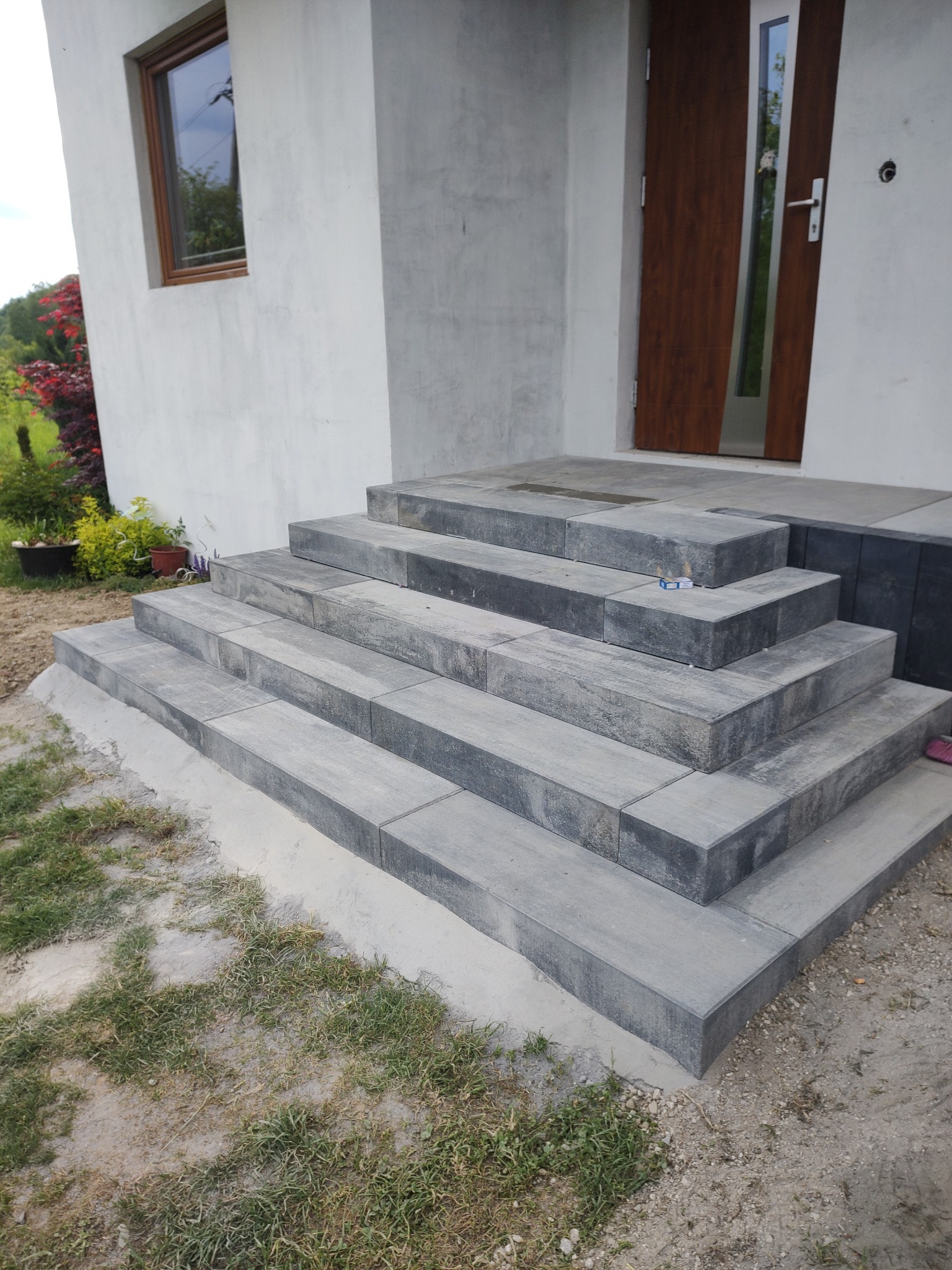 Schody wejściowe do domu wyłożone szarą kostką brukową, z pudełkiem zapałek leżącym na drugim stopniu, widoczne rośliny w doniczkach i brązowe drzwi wejściowe.