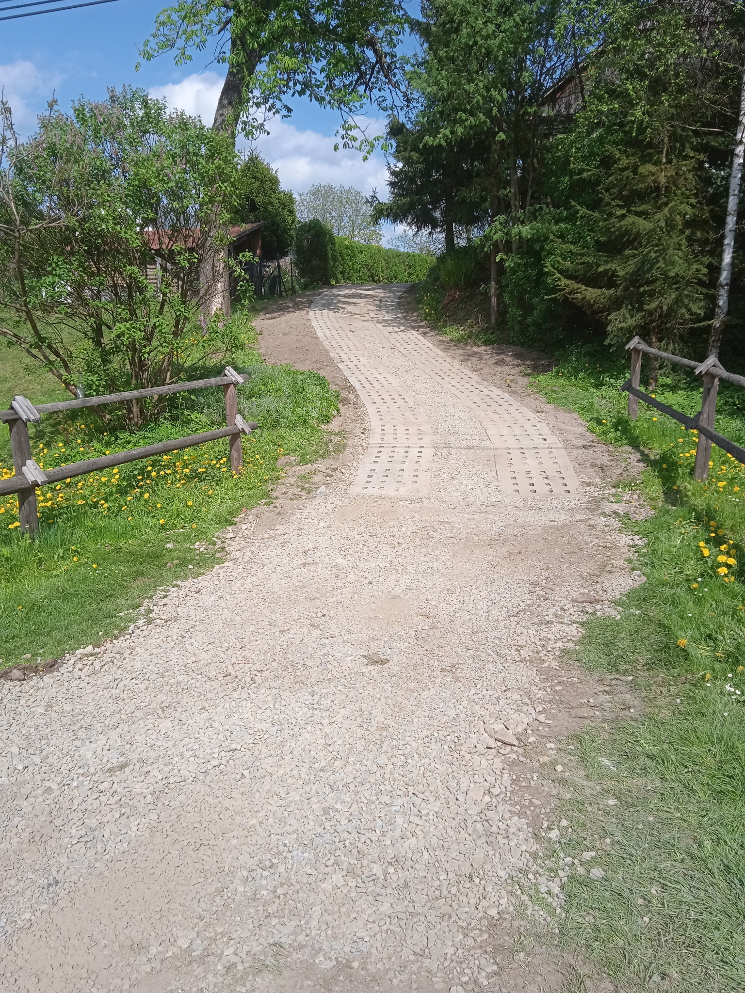 Ujęcie drogi gruntowej z fragmentami utwardzonej nawierzchni z ażurowych płyt betonowych, otoczonej zielenią i drewnianym płotkiem, prowadzącej pod górę w kierunku zabudowań.