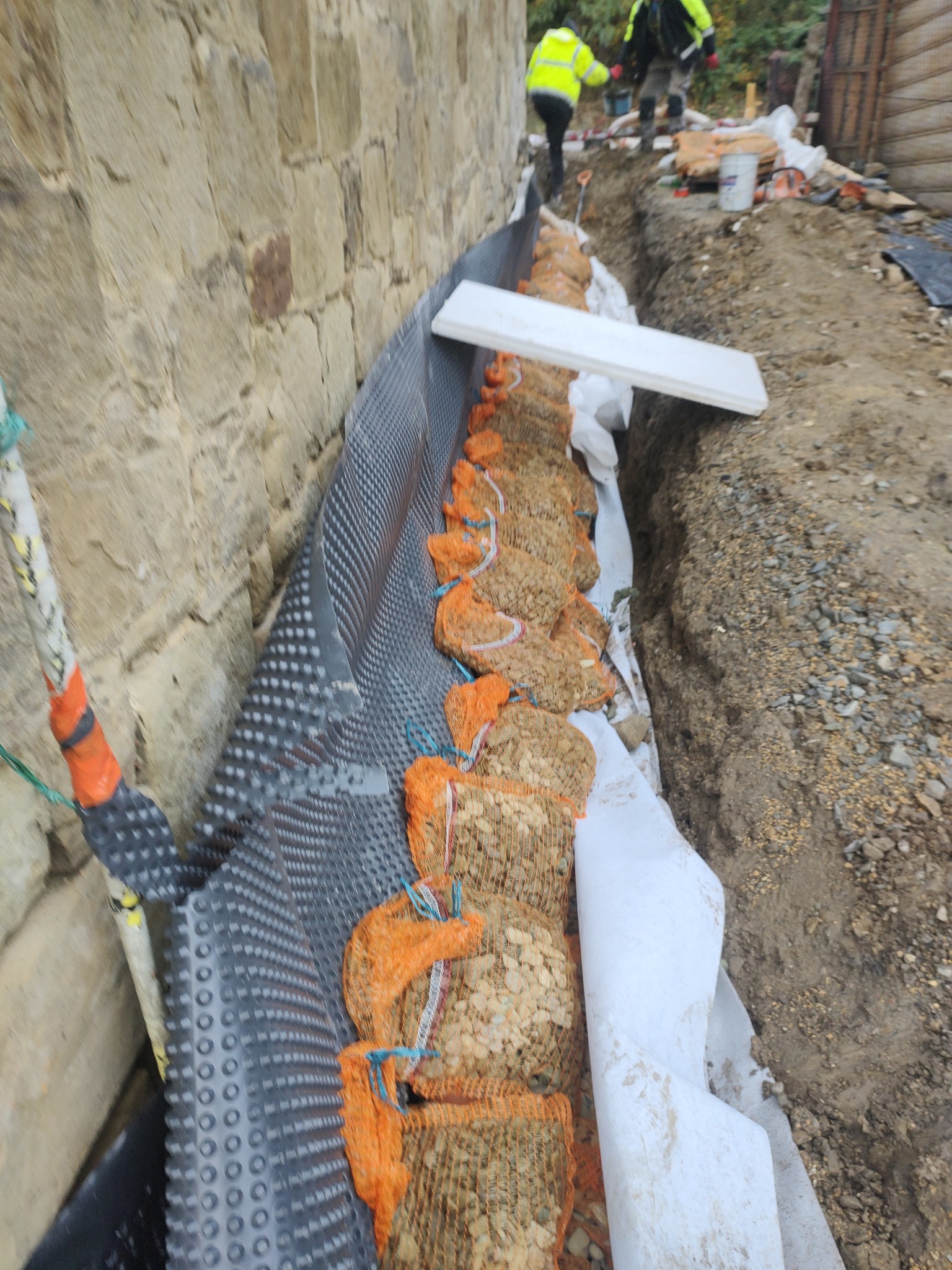 Ocieplenie i wzmocnienie kamiennej ławicy fundamentowej. Widoczna folia kubełkowa, geowłóknina i worki z kamieniem. W tle pracownicy w odblaskowych kamizelkach.