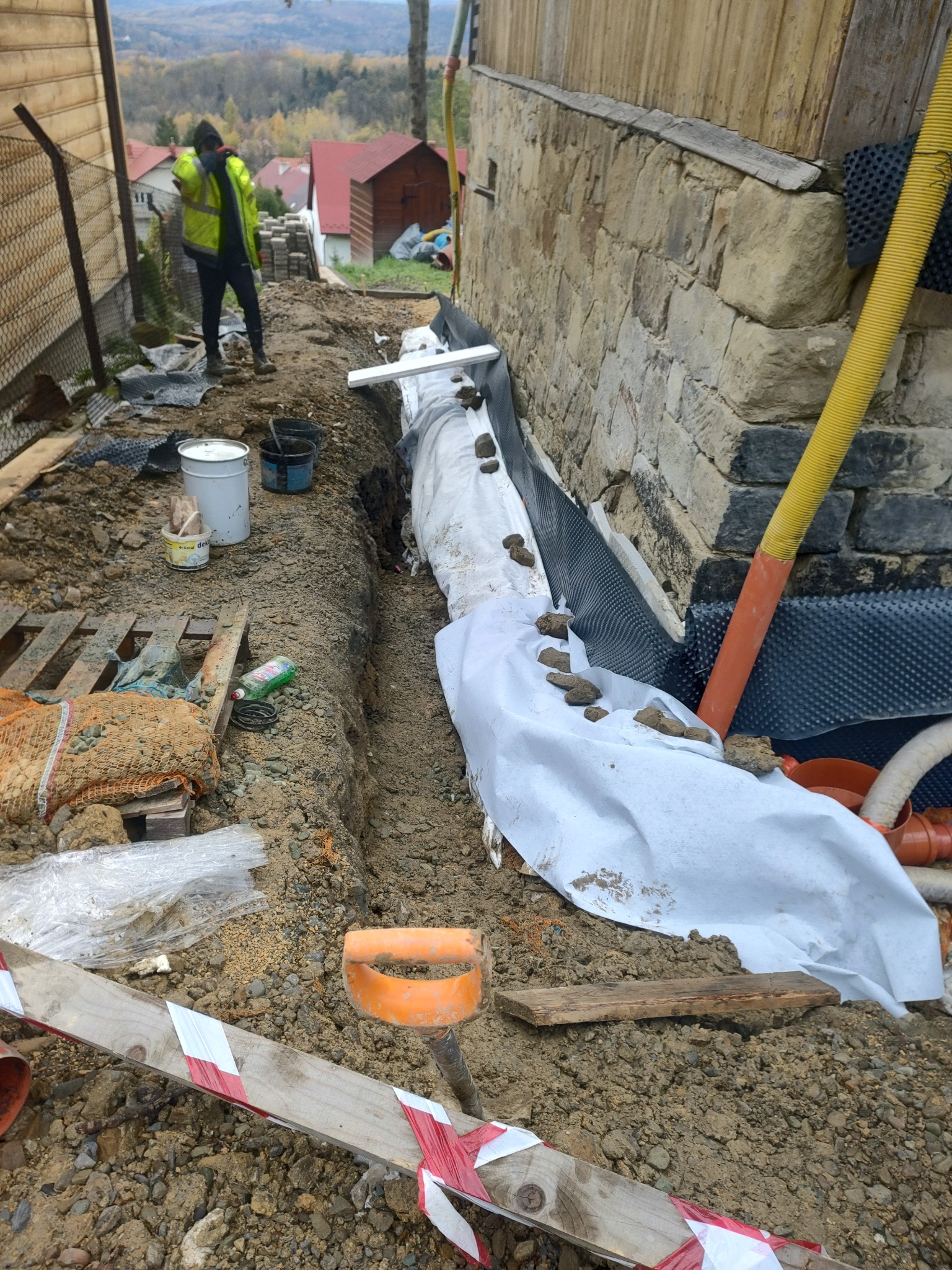 Wykop w ziemi wzdłuż kamiennej ściany fundamentowej, zabezpieczony geowłókniną i folią kubełkową, z widocznymi rurami drenażowymi i pracownikiem w odblaskowej kamizelce w tle.