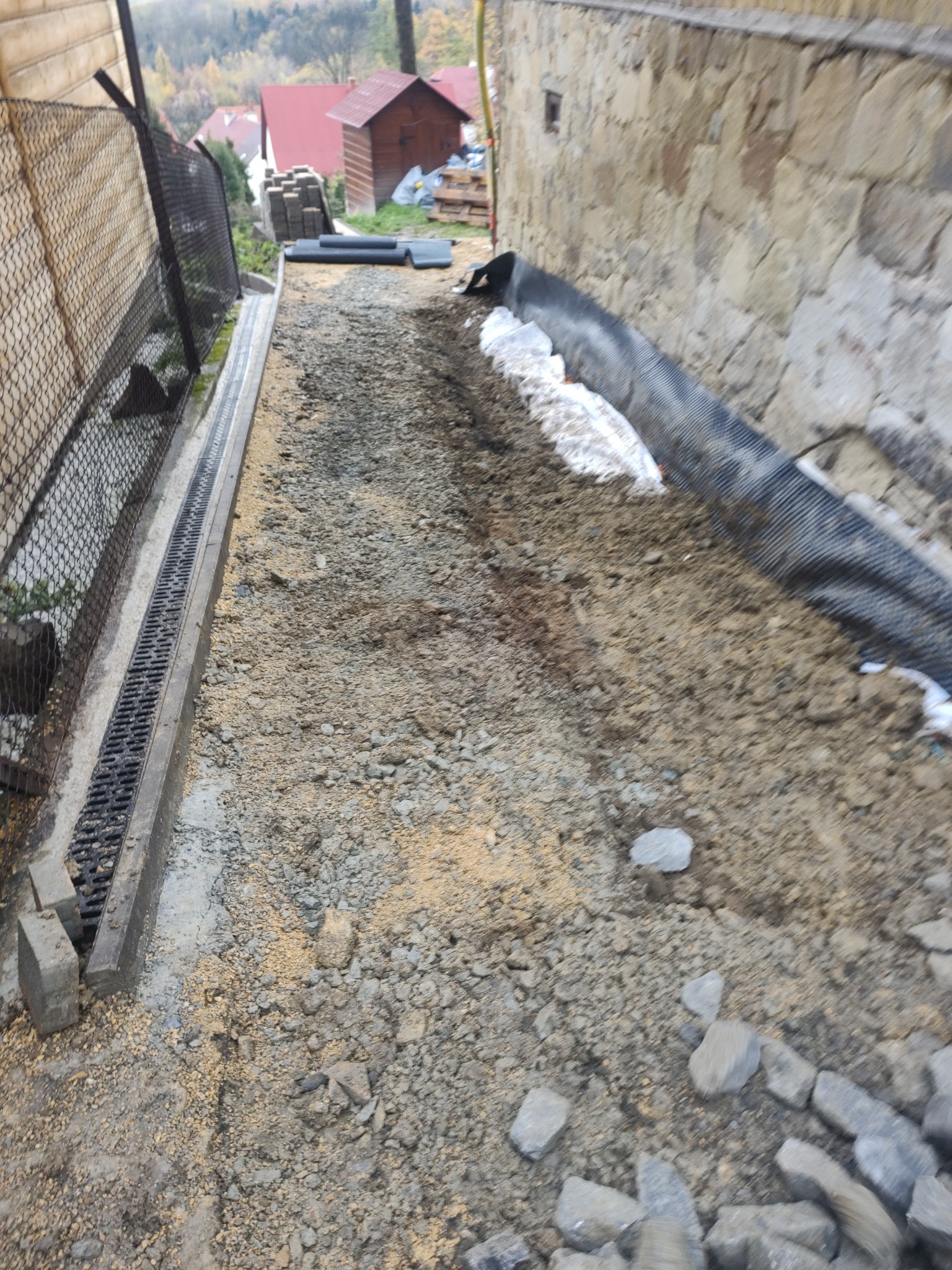 Przygotowanie terenu pod budowę: drenaż liniowy z kratką, warstwa gruntu z drobnym kruszywem, geowłóknina przy kamiennej ścianie, widoczne elementy drenażu i kamienie.