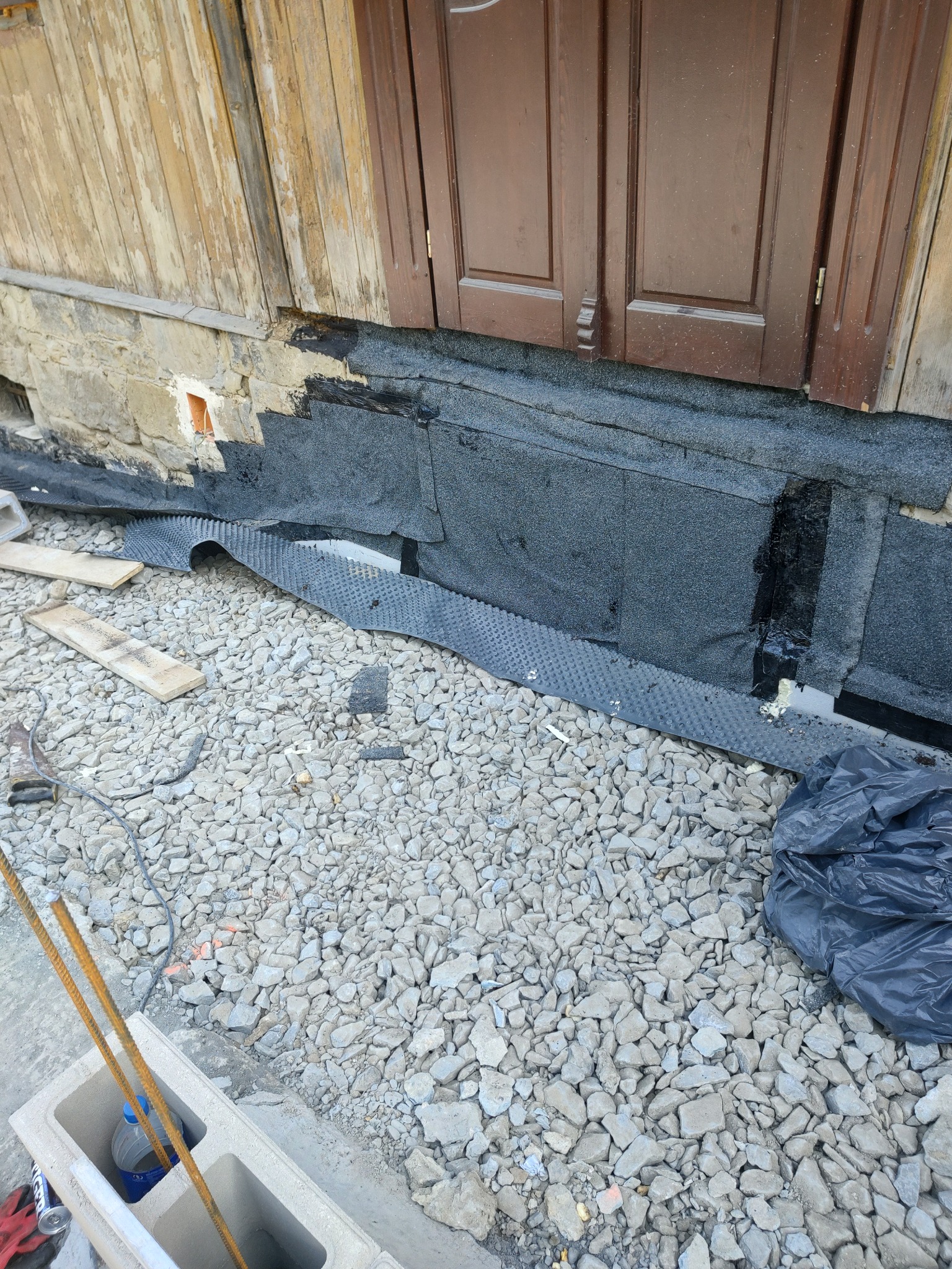 Ocieplenie kamiennej ławicy fundamentowej z użyciem hydroizolacji bitumicznej i folii kubełkowej, widoczne wzmocnienie betonowe, gruz i elementy konstrukcyjne w trakcie prac.