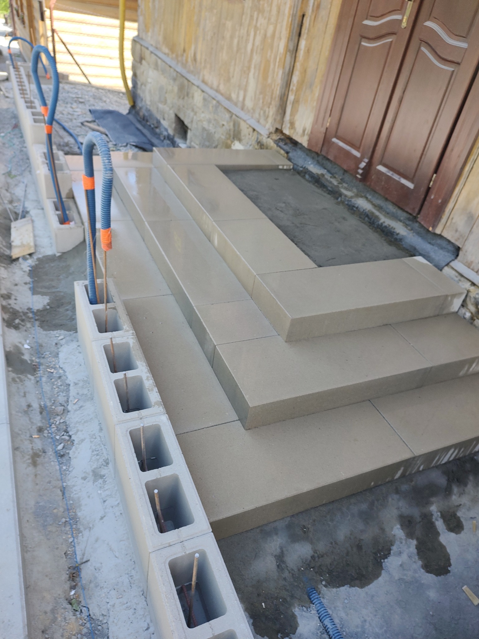 Schody wejściowe wyłożone jasnymi płytami kamiennymi, obok wzniesiona ściana z betonowych pustaków z wystającymi prętami zbrojeniowymi i peszlami instalacyjnymi, widok z góry.