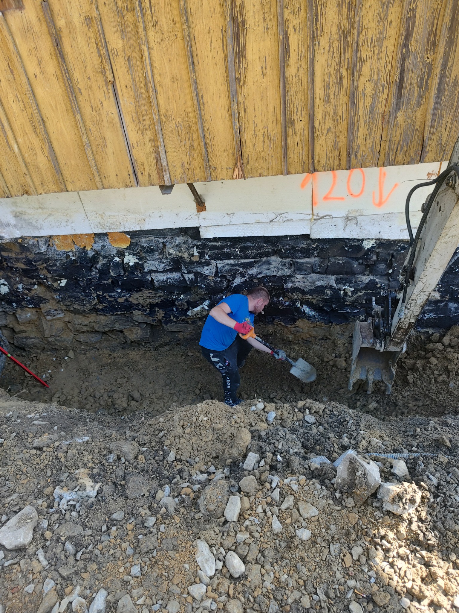 Pracownik w niebieskiej koszulce kopie łopatą dół obok fundamentu budynku, z widocznym fragmentem koparki i oznaczeniem '120' na ścianie.