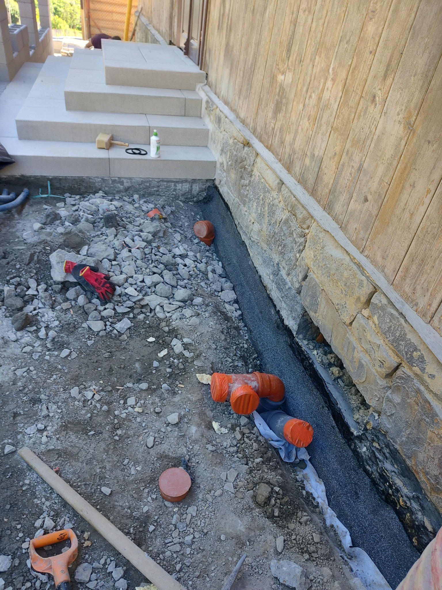 Przygotowanie podłoża pod schody zewnętrzne: widoczne warstwy izolacyjne, drenaż z rur kanalizacyjnych, kamienna ławica fundamentowa oraz narzędzia pracy.