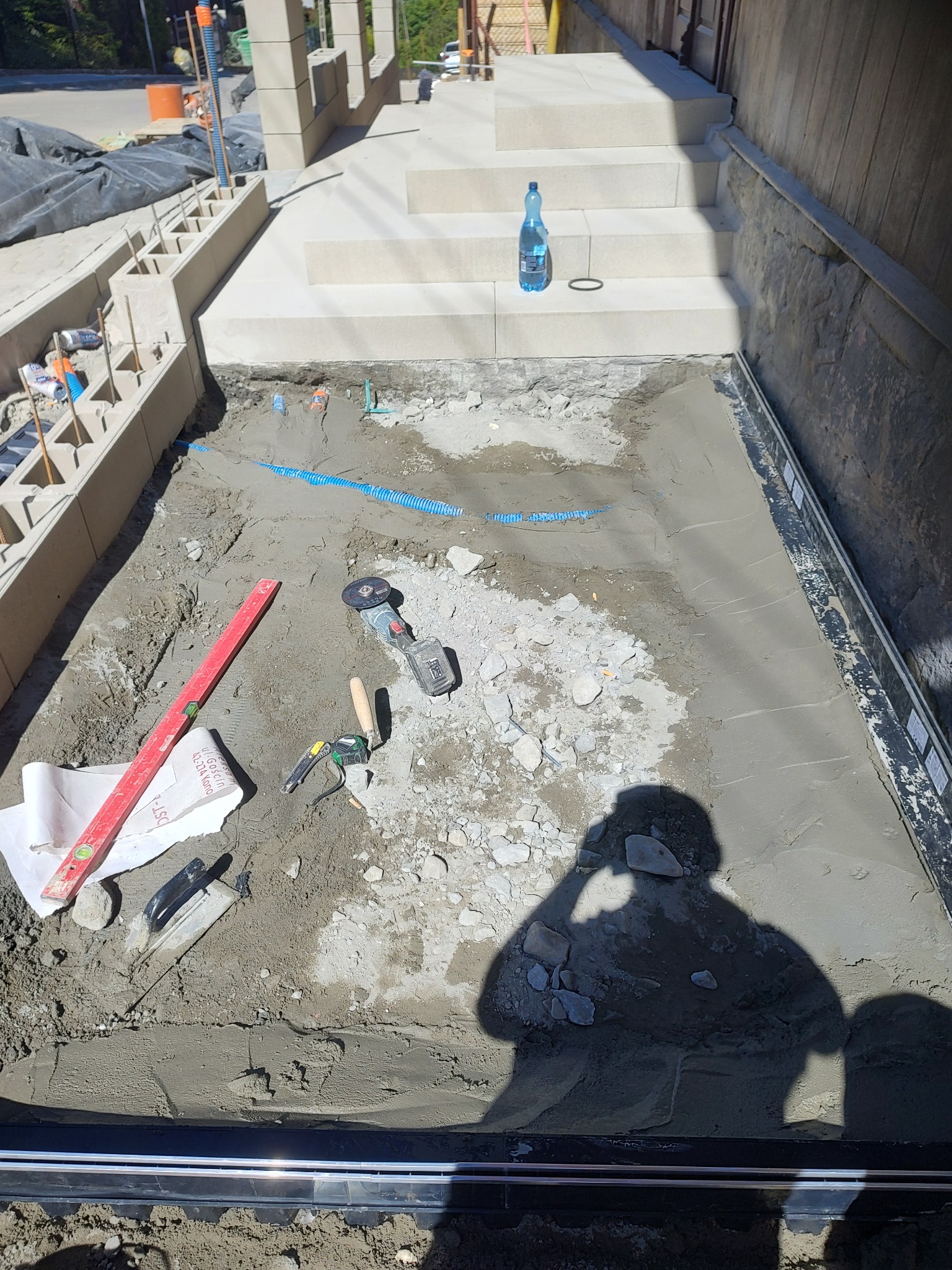Przygotowanie podłoża pod schody zewnętrzne: wylany beton z widocznymi niebieskimi rurami, poziomica, kielnia i szlifierka kątowa na powierzchni, cień osoby robiącej zdjęcie.