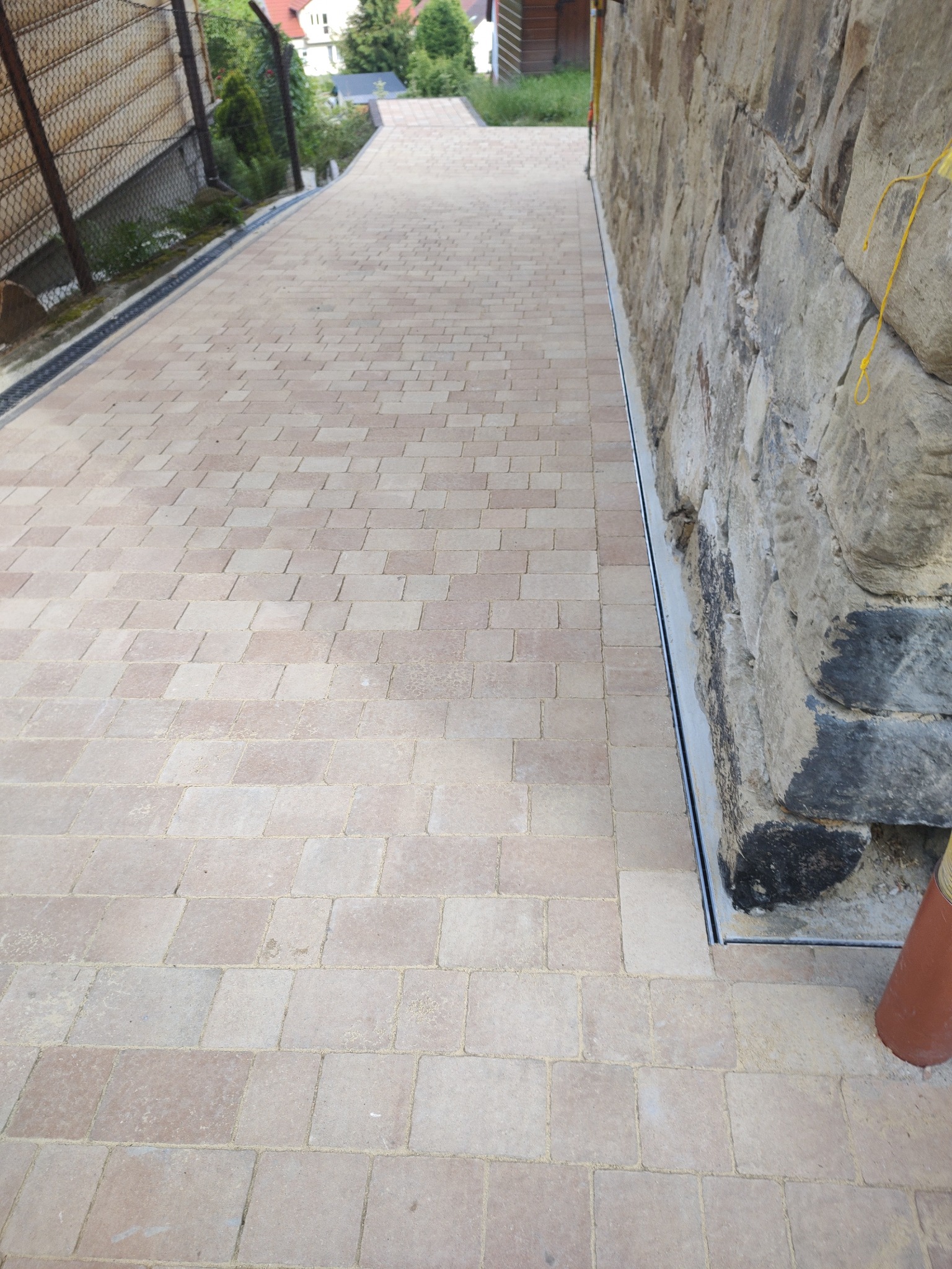 Ułożona kostka brukowa w odcieniach beżu i brązu, przylegająca do kamiennej ściany z zamontowanym odpływem liniowym, widok z góry.