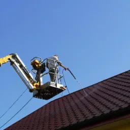 Pracownik na podnośniku koszowym czyści dach z metalowej dachówki przy użyciu myjki ciśnieniowej, widok na błękitne niebo.