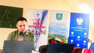 Mężczyzna prowadzi prelekcję o ratownictwie, za nim banery: 'Zostań Ratownikiem!', herb powiatu kieleckiego i logo Państwowej Straży Pożarnej. Laptop na stole.