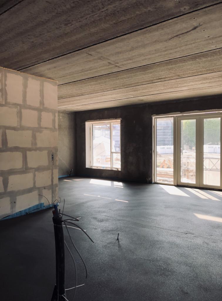 Świeżo wylana, ciemna posadzka w surowym wnętrzu z oknami i widoczną konstrukcją budynku w tle, z widocznym sprzętem do wylewania w rogu.