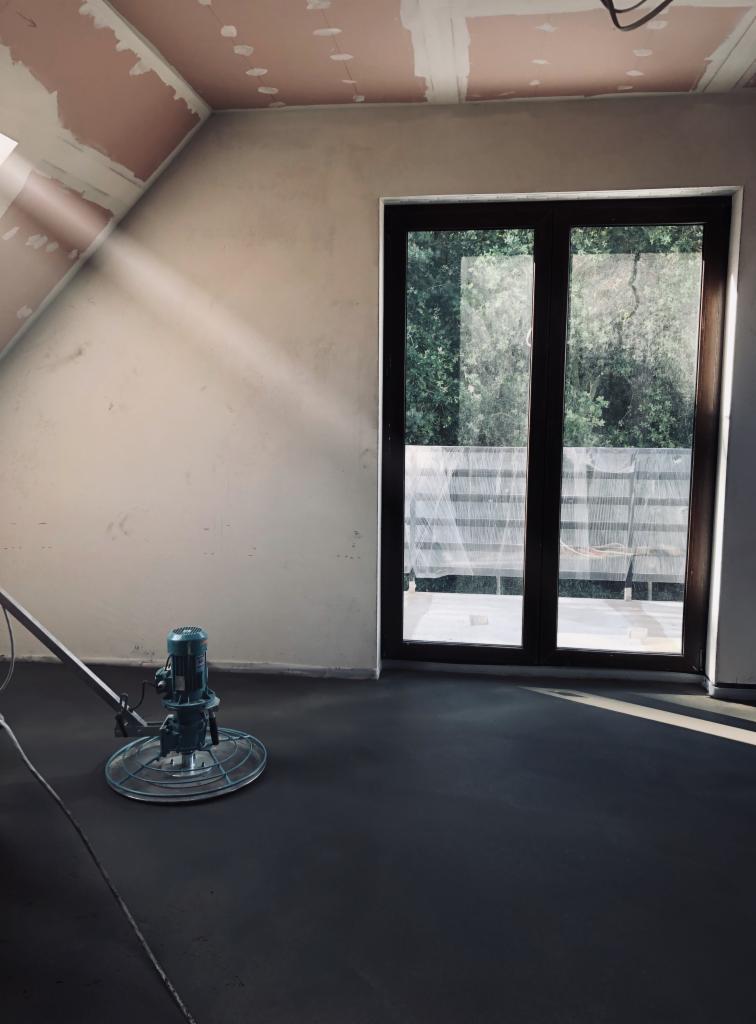 Szlifowanie mokrego, ciemnoszarego jastrychu maszyną w pomieszczeniu z dużym oknem balkonowym i surowymi ścianami pod skosem, promień światła wpadający przez okno.