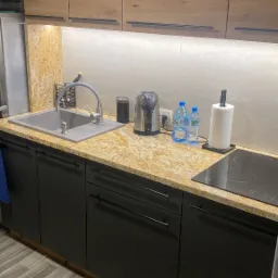 Fragment nowoczesnej kuchni z blatem o złocistej fakturze, czarnymi szafkami z minimalistycznymi uchwytami, szarym zlewozmywakiem i płytą indukcyjną. Na blacie czajnik elektryczny, dozownik...