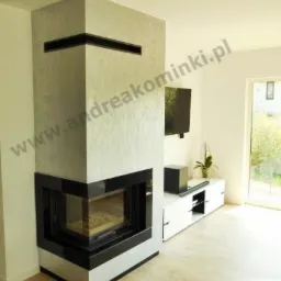Nowoczesny kominek narożny z obudową w odcieniach szarości i czarnymi elementami wykończeniowymi, widoczny w jasnym salonie z telewizorem i białymi meblami.