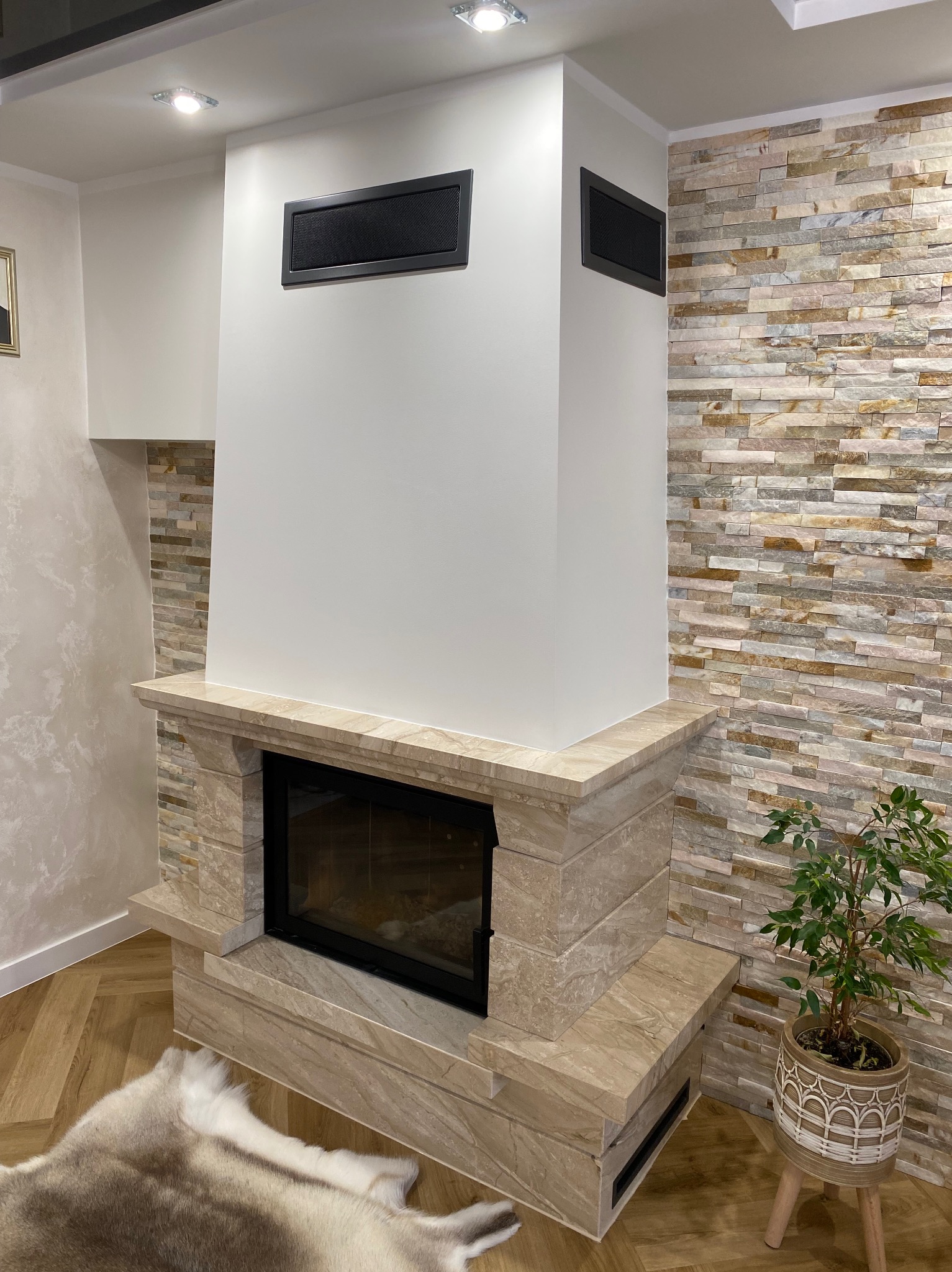 Nowoczesny kominek z obudową z jasnego kamienia i białym wykończeniem, uzupełniony dekoracyjną ścianą z kamienia łupanego i rośliną doniczkową.