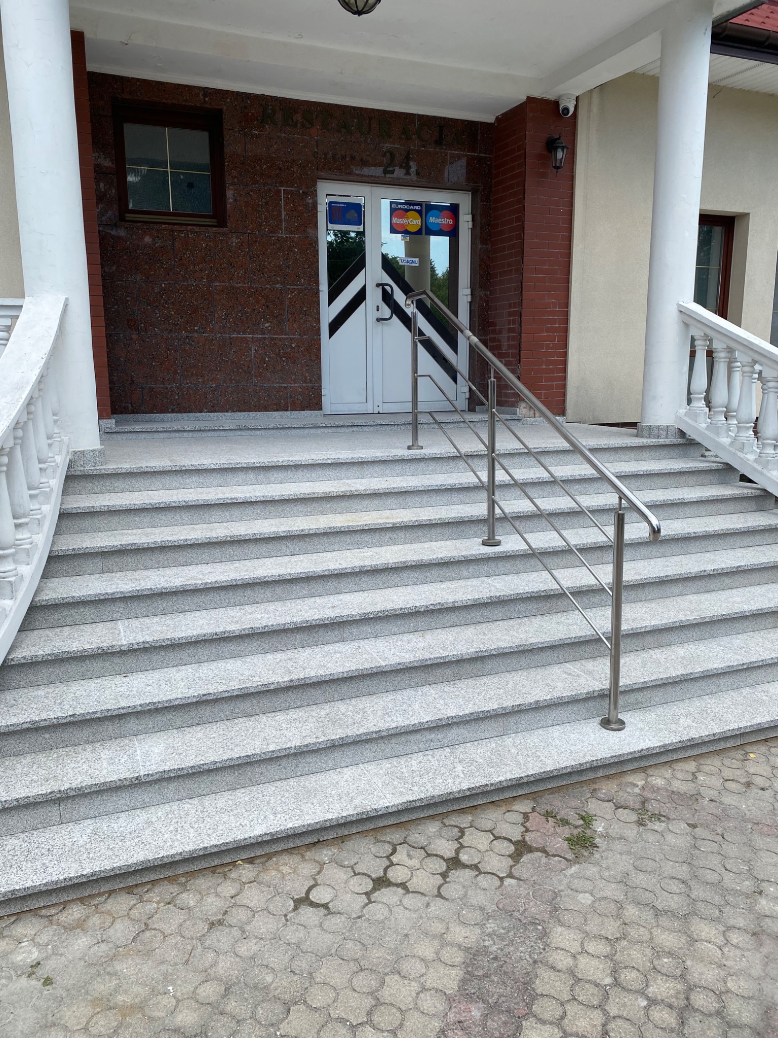 Zewnętrzne schody wejściowe wyłożone szarym kamieniem z metalową balustradą, prowadzące do budynku z kolumnami i drzwiami z naklejkami kart płatniczych.