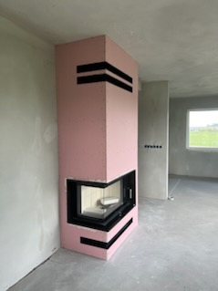 Surowy kominek narożny w trakcie budowy, obłożony różowym materiałem izolacyjnym z czarnymi, poziomymi pasami, w stanie deweloperskim z widocznym oknem i polami w tle.