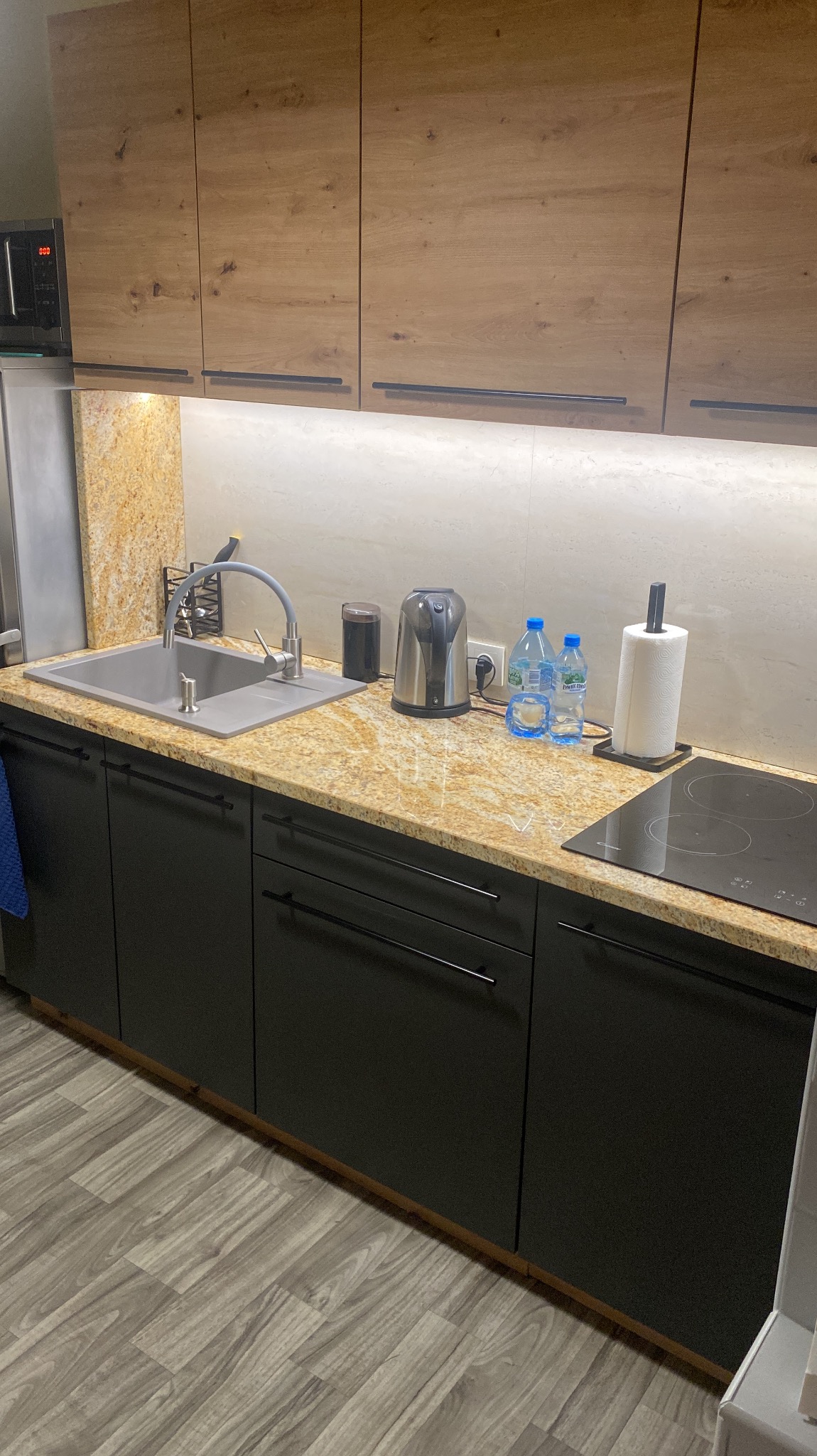 Fragment nowoczesnej kuchni z blatem o złocistej fakturze, czarnymi szafkami z minimalistycznymi uchwytami, szarym zlewozmywakiem i płytą indukcyjną. Na blacie czajnik elektryczny, dozownik...