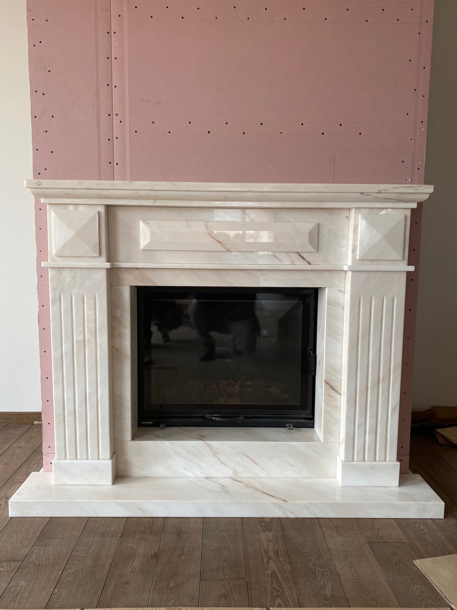 Marmurowa obudowa kominka z czarnym wkładem, widoczna na tle ściany z płyt kartonowo-gipsowych w trakcie montażu.