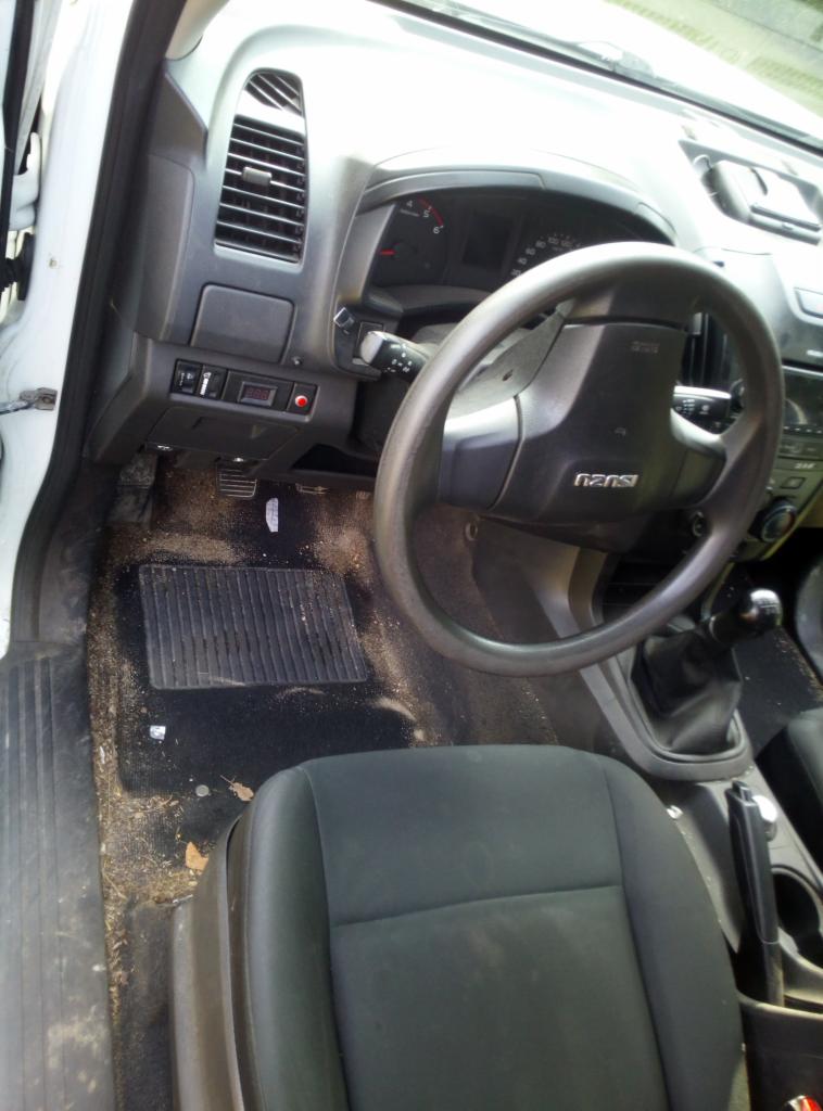 Wnętrze samochodu marki Isuzu z widocznymi zabrudzeniami na dywaniku i fotelu kierowcy, deska rozdzielcza z kierownicą i elementami sterującymi.