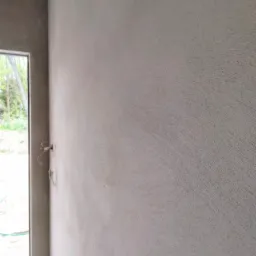 Świeżo nałożony, surowy tynk maszynowy na ścianie wewnętrznej, widoczne przewody elektryczne wystające ze ściany, krawędź okna z widokiem na zieleń na zewnątrz.