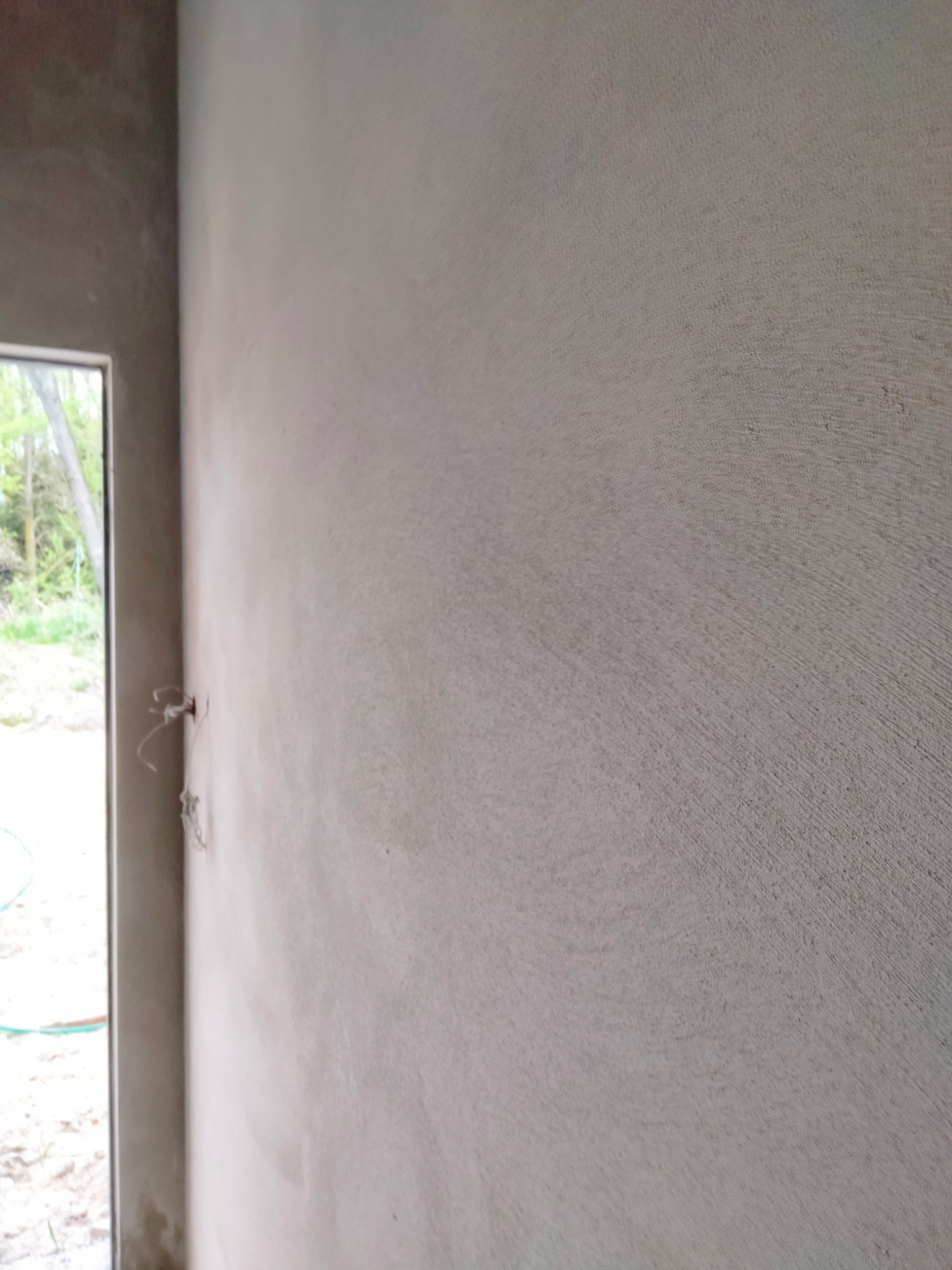 Świeżo nałożony, surowy tynk maszynowy na ścianie wewnętrznej, widoczne przewody elektryczne wystające ze ściany, krawędź okna z widokiem na zieleń na zewnątrz.