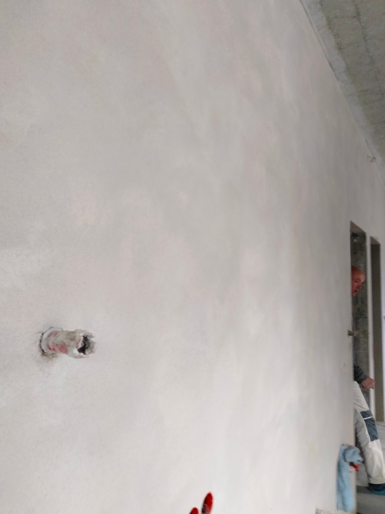 Świeżo nałożony, jasnoszary tynk na ścianie wewnętrznej z widocznymi śladami po szpachli, z wystającą rurą instalacyjną i fragmentem sylwetki pracownika w tle.