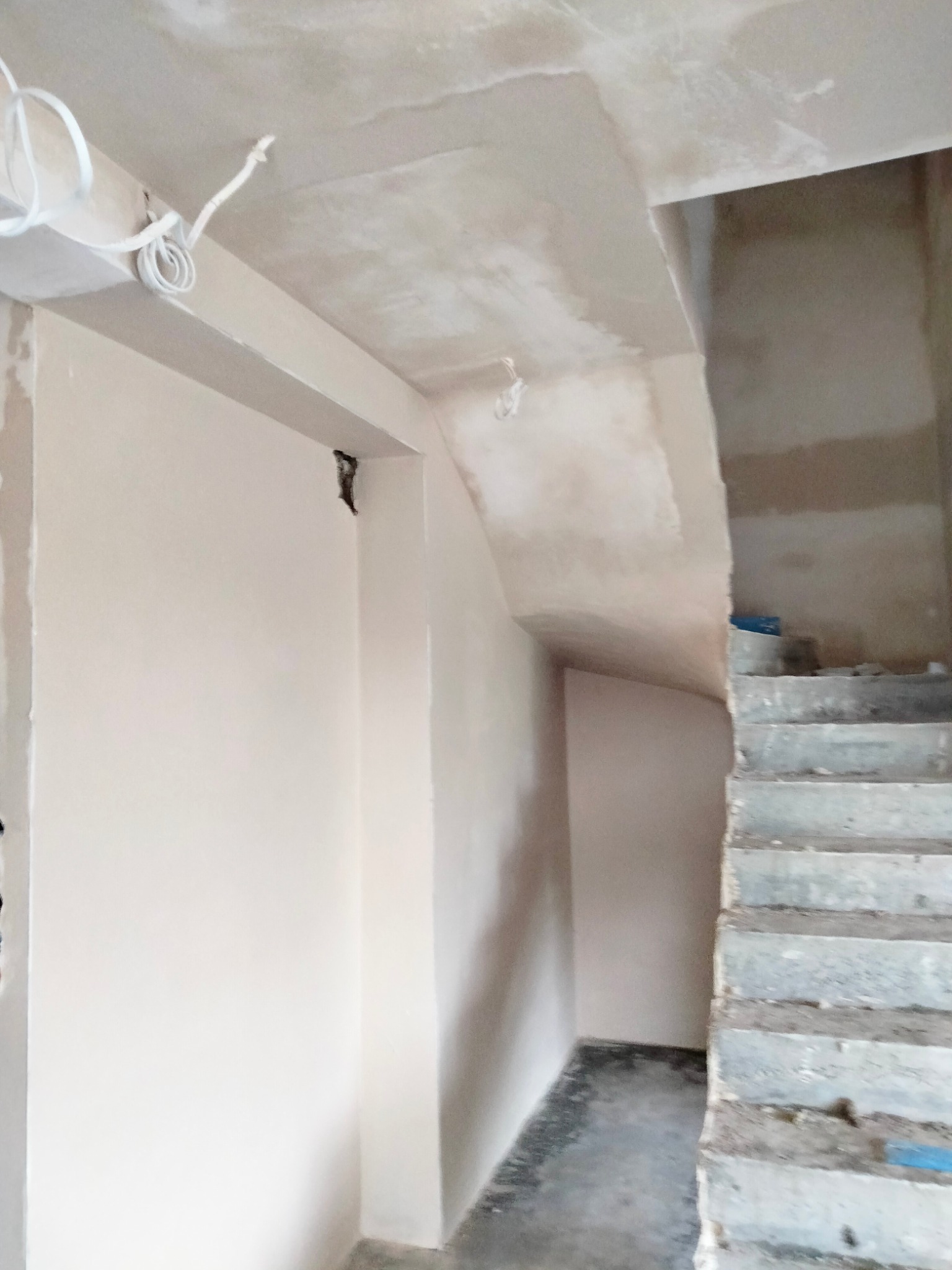 Świeżo otynkowana ściana w jasnym odcieniu z widocznymi przewodami elektrycznymi pod sufitem oraz surowymi schodami w tle, ukazująca wnętrze w trakcie prac wykończeniowych.