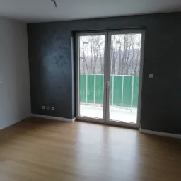 Pusty pokój po remoncie z panelami podłogowymi w odcieniach brązu, ściana pomalowana na ciemny szary z efektem dekoracyjnym, widoczne gniazdka elektryczne i wystające kable z sufitu.